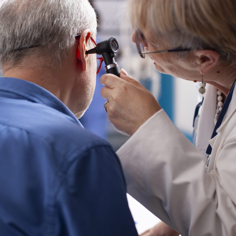 Experienced retired female physician inspecting ear of elderly male patient in clinic office. Closeup of skilled old doctor using otoscope during medical consultation of senior white man.